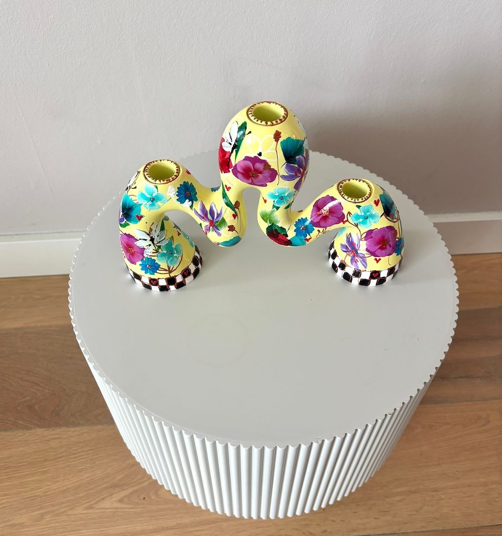 Een gele, golvende kandelaar met bloemmotieven en een zwart-wit geblokte voet op een witte ronde tafel.