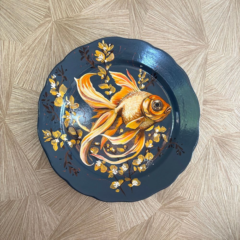 Decoratief bord met een geschilderde goudvis op een donkerblauwe achtergrond, op een beige, gestructureerde ondergrond.