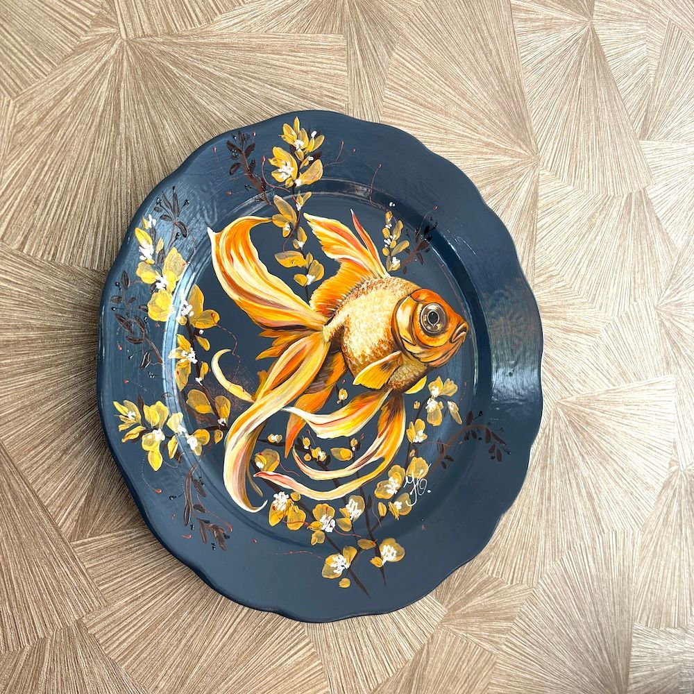 Decoratief bord met een geschilderde goudvis op een donkerblauwe achtergrond, op een beige, gestructureerde ondergrond.