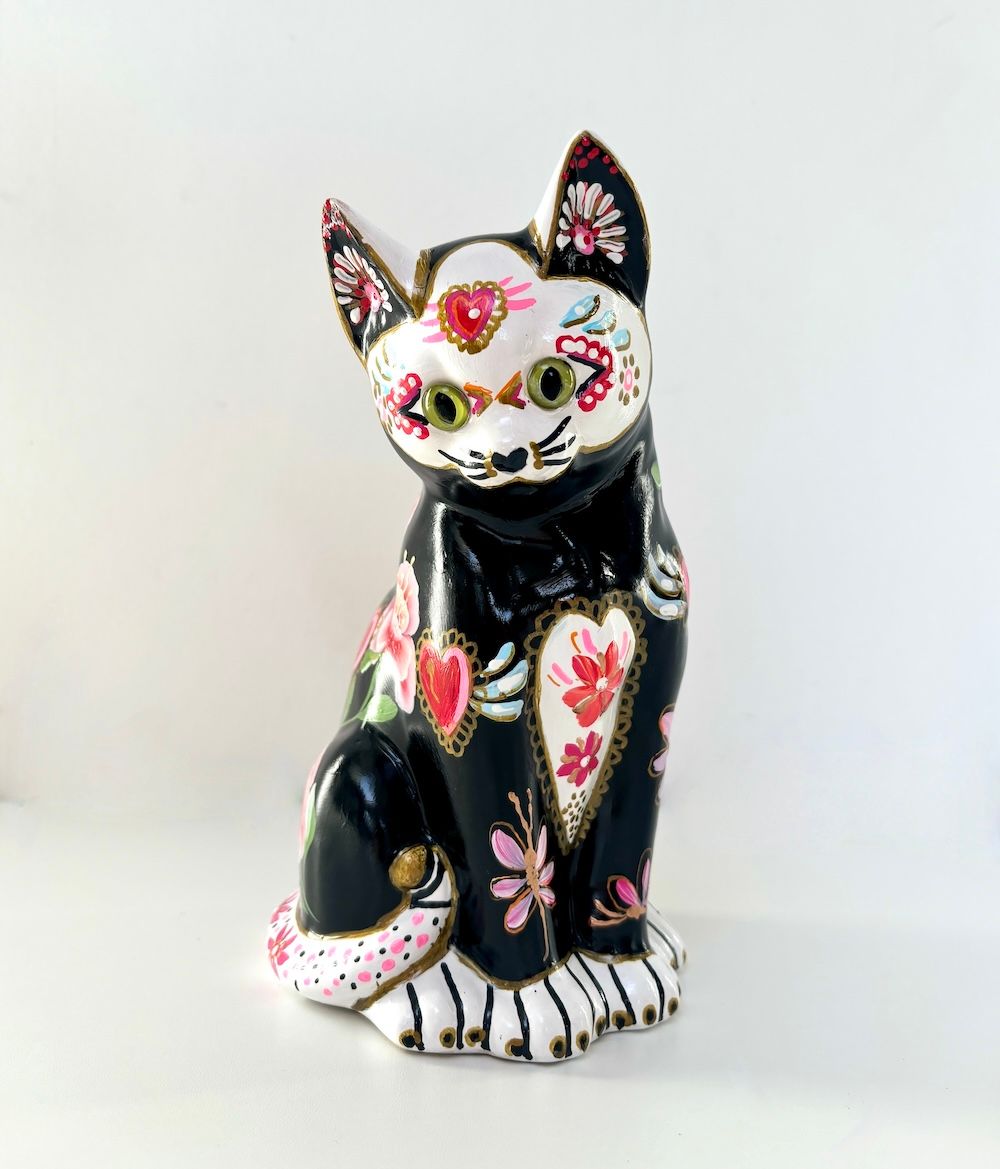 Een beeldje van een zwarte kat, versierd in de stijl van Día de Muertos ,waaronder roze hartjes, bloemen en witte details.