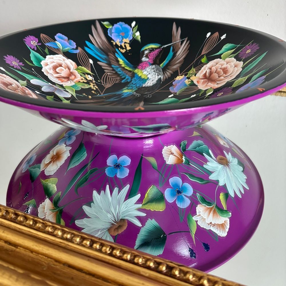 Zwarte bloemenschaal met een kleurrijke vogel, rustend op een paarse bloemenbasis in een gouden frame.