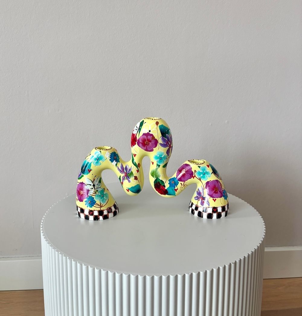 Een gele, golvende kandelaar met bloemmotieven en een zwart-wit geblokte voet op een witte ronde tafel.