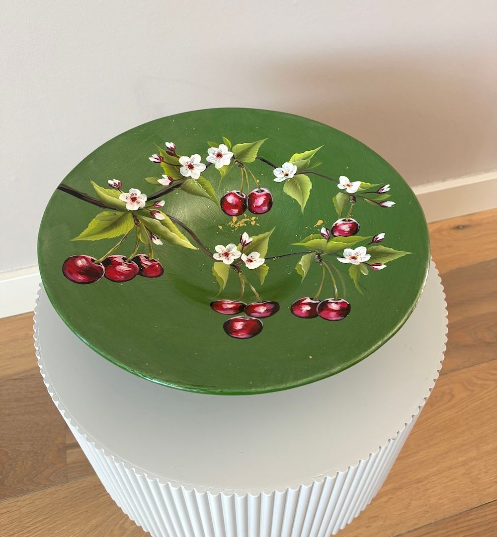 Een ronde, groene, handbeschilderde schaal met kersenbloesems en rode kersen staat op een witte, geplooide ronde tafel.