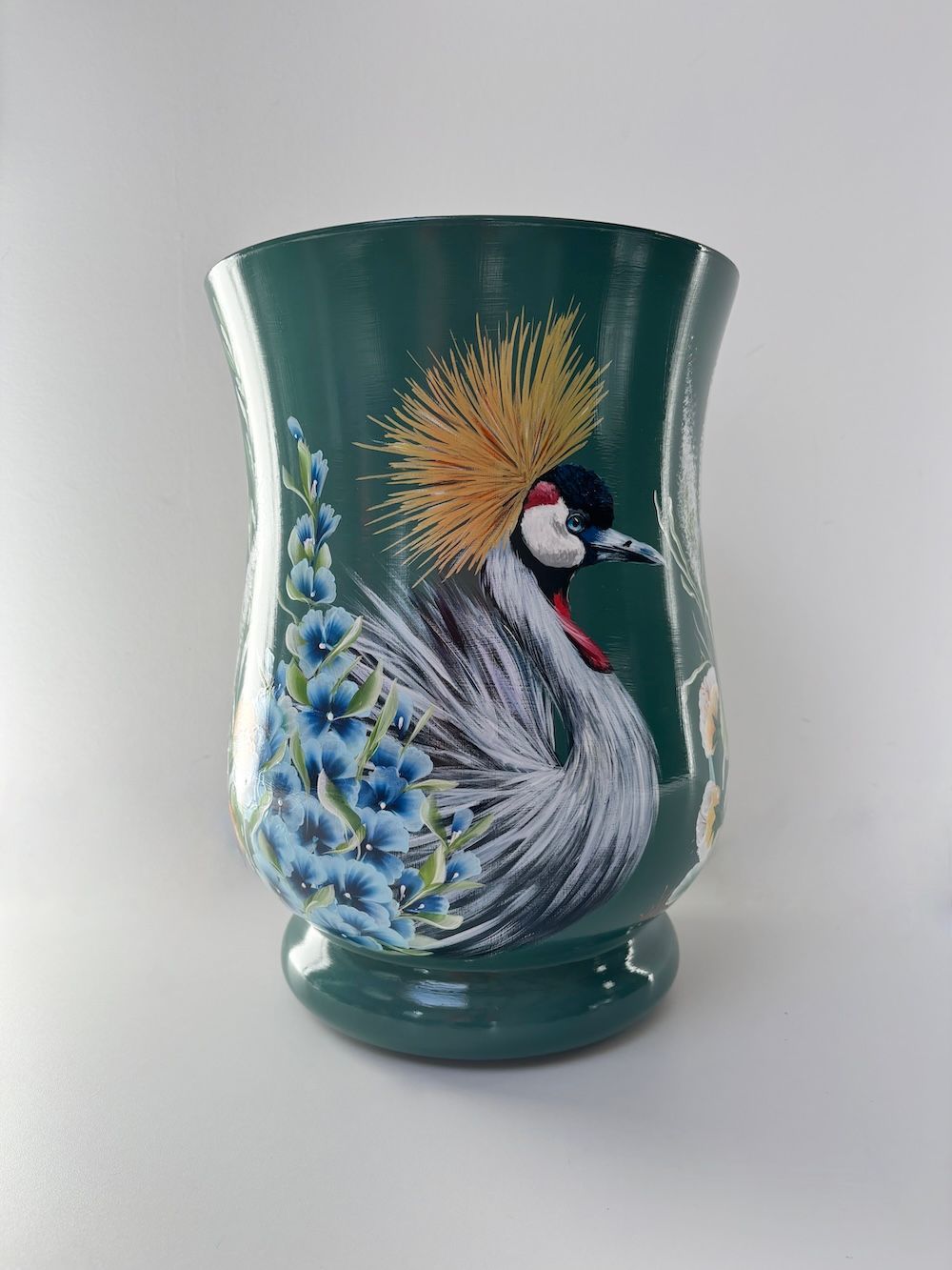 Een donkergroene vaas met een geschilderde grijze kraanvogel met een gele kuif, die naast een blauw bloemstuk staat.