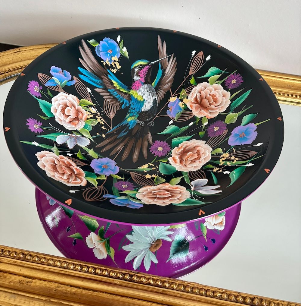 Zwarte bloemenschaal met een kleurrijke vogel, rustend op een paarse bloemenbasis in een gouden frame.