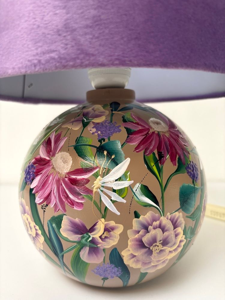 Een lamp met een ronde keramische voet waarop bloemen zijn geschilderd, en een effen paarse fluwelen lampenkap.