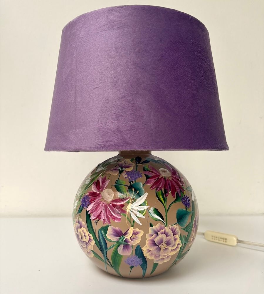 Een lamp met een ronde keramische voet waarop bloemen zijn geschilderd, en een effen paarse fluwelen lampenkap.