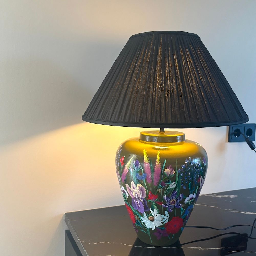 Decoratieve tafellamp met bloemmotief en zwarte geplooide kap op een donkere tafel tegen de muur.