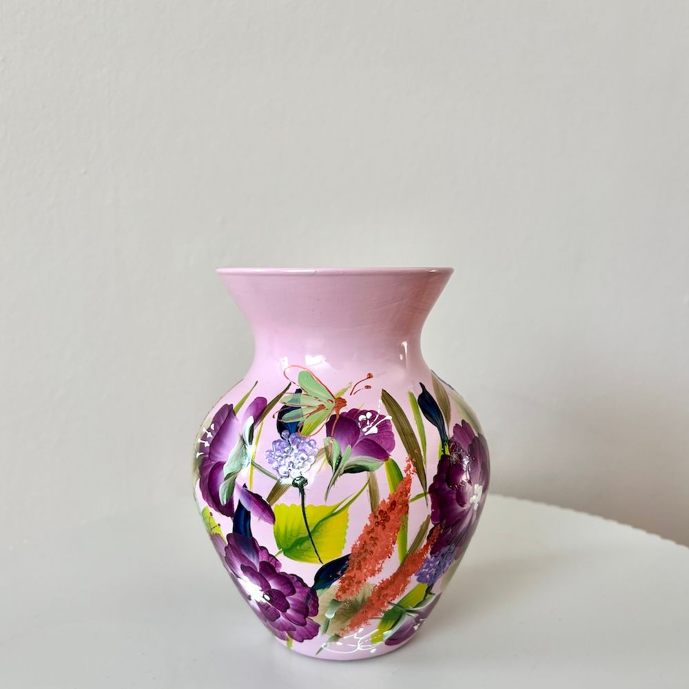 Een roze, uitlopende keramische vaas, beschilderd met een patroon van paarse bloemen, groene bladeren en oranje tarwehalmen.