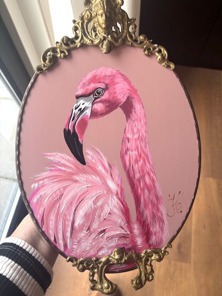 Een schilderij van een roze flamingo in een sierlijke, ovale gouden lijst aan een lichte muur.