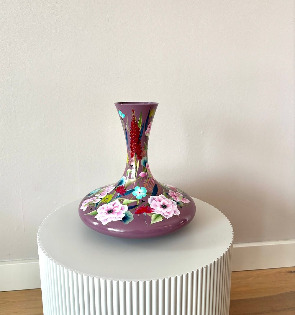 Paarse glazen vaas met handgeschilderde roze en blauwe bloemen op een witte tafel.