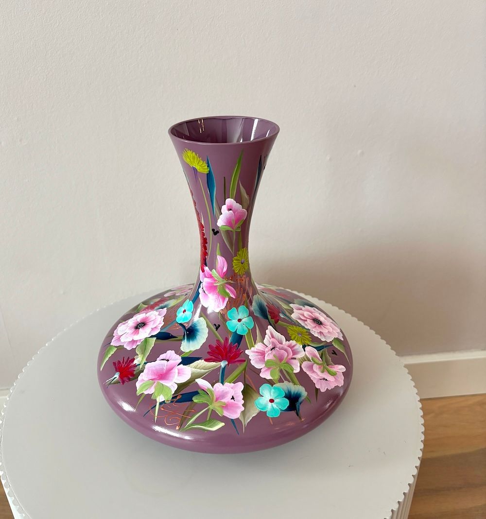 Paarse glazen vaas met handgeschilderde roze en blauwe bloemen op een witte tafel.