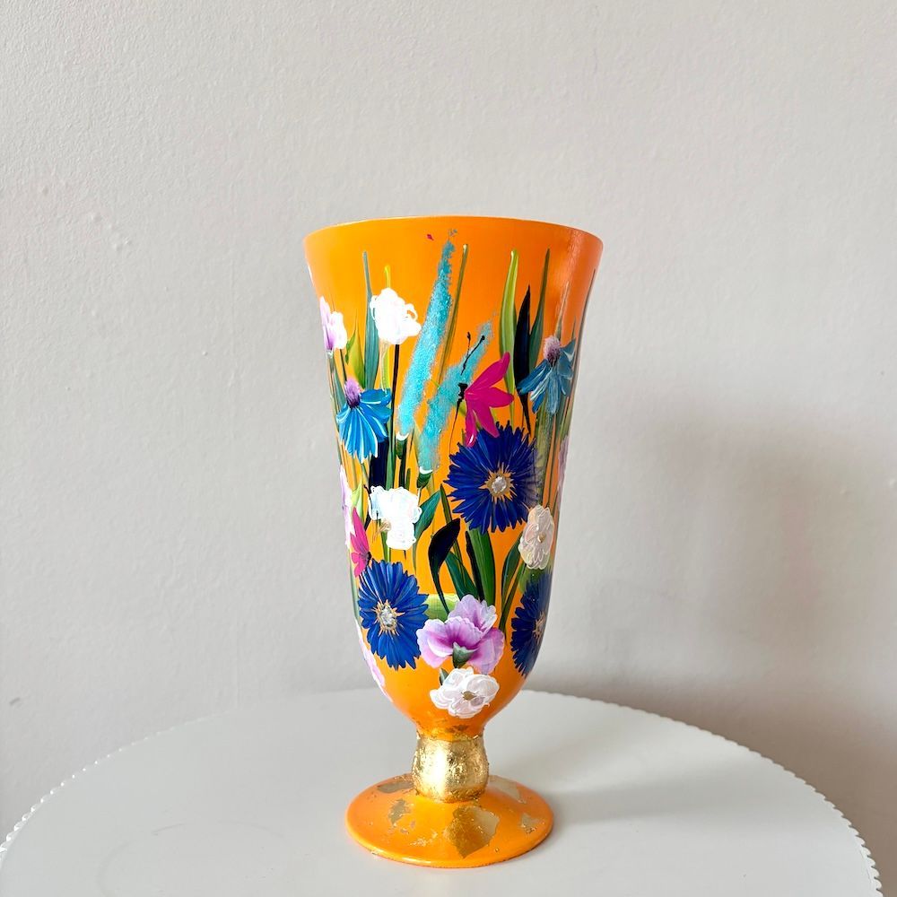 Een hoge, oranje vaas op voet, beschilderd met kleurrijke bloemmotieven en groen bladwerk, met gouden accenten op de voet.