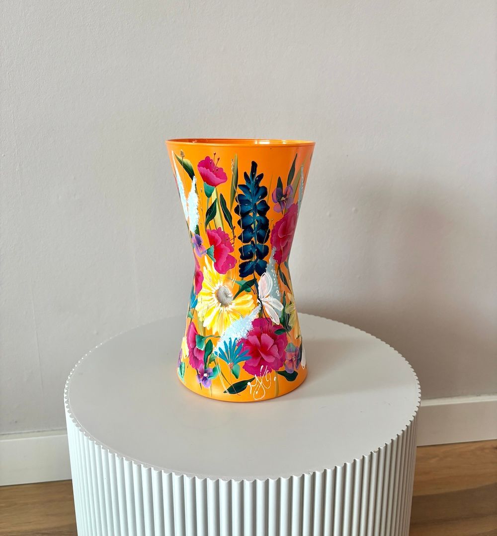 Een handbeschilderde oranje vaas met levendige, kleurrijke bloemmotieven staat centraal op een witte, gegroefde sokkel.