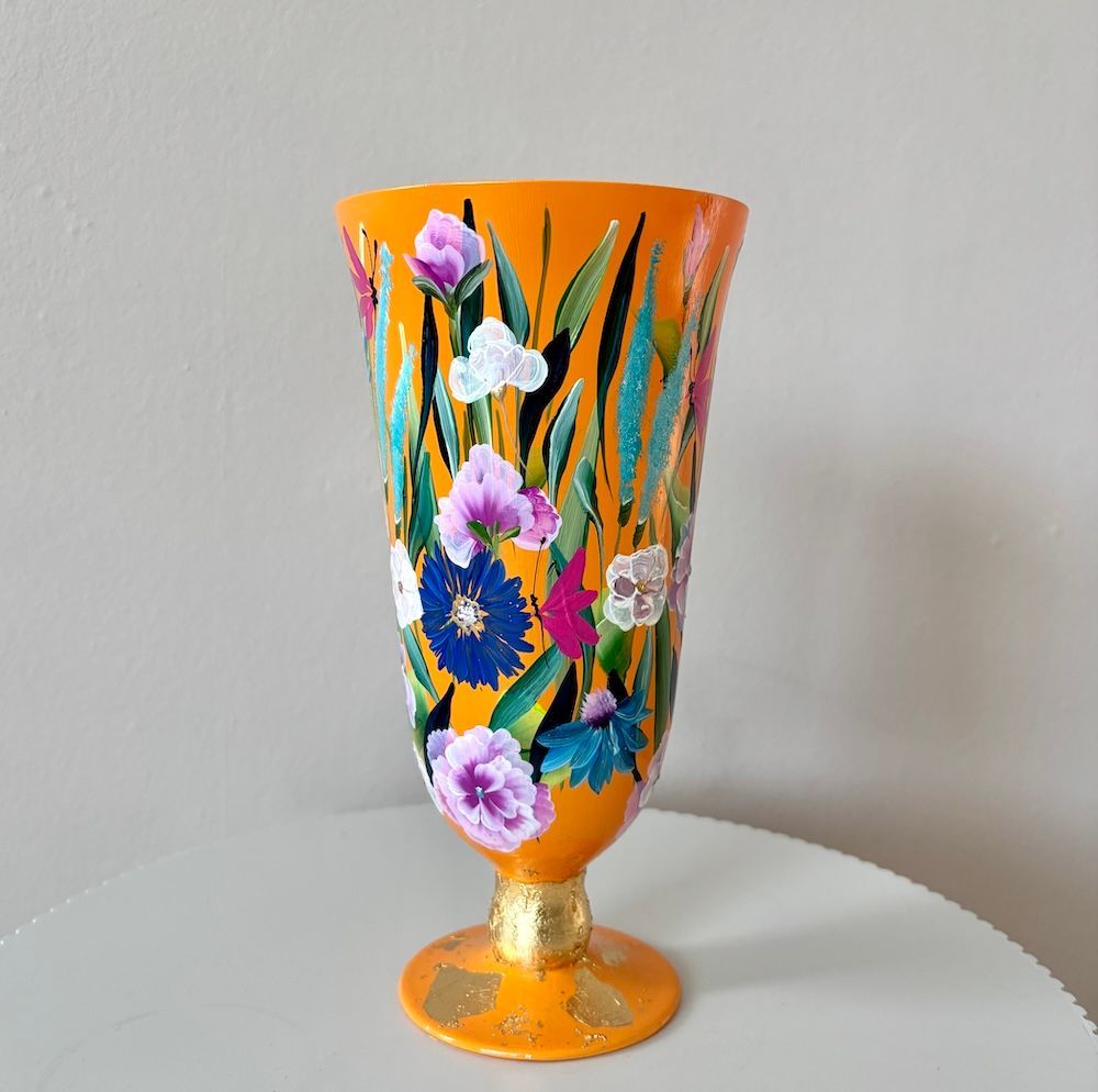 Een hoge, oranje vaas op voet, beschilderd met kleurrijke bloemmotieven en groen bladwerk, met gouden accenten op de voet.