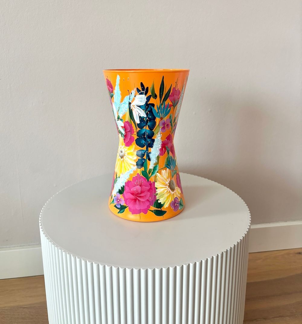 Een handbeschilderde oranje vaas met levendige, kleurrijke bloemmotieven staat centraal op een witte, gegroefde sokkel.