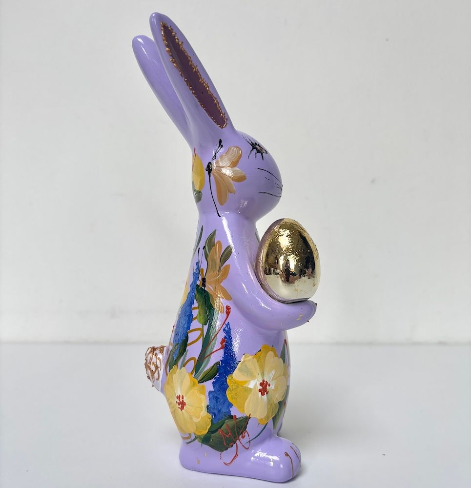 Paars decoratief konijntje dat een gouden ei vasthoudt, met een lijfje beschilderd met bloemen op een witte achtergrond.