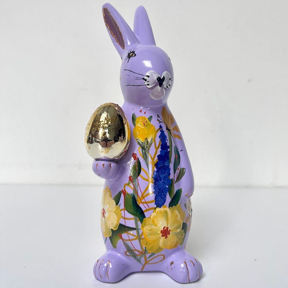 Paars decoratief konijntje dat een gouden ei vasthoudt, met een lijfje beschilderd met bloemen op een witte achtergrond.