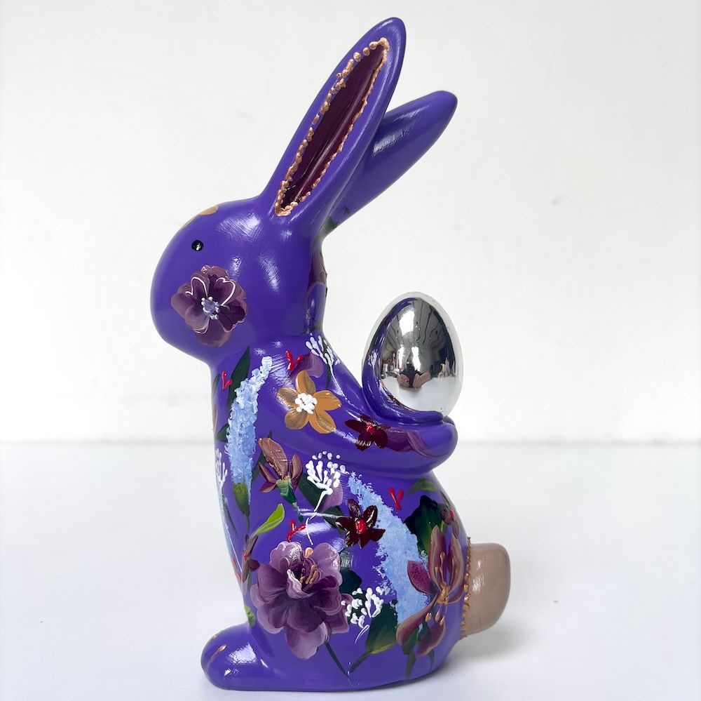 Paarse konijnenfiguur met bloemenmotief die een zilveren eivormig ornament vasthoudt.