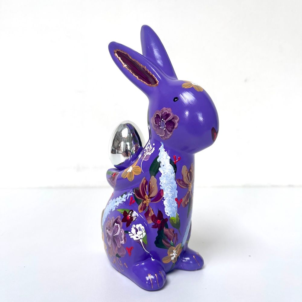 Paarse konijnenfiguur met bloemenmotief die een zilveren eivormig ornament vasthoudt.