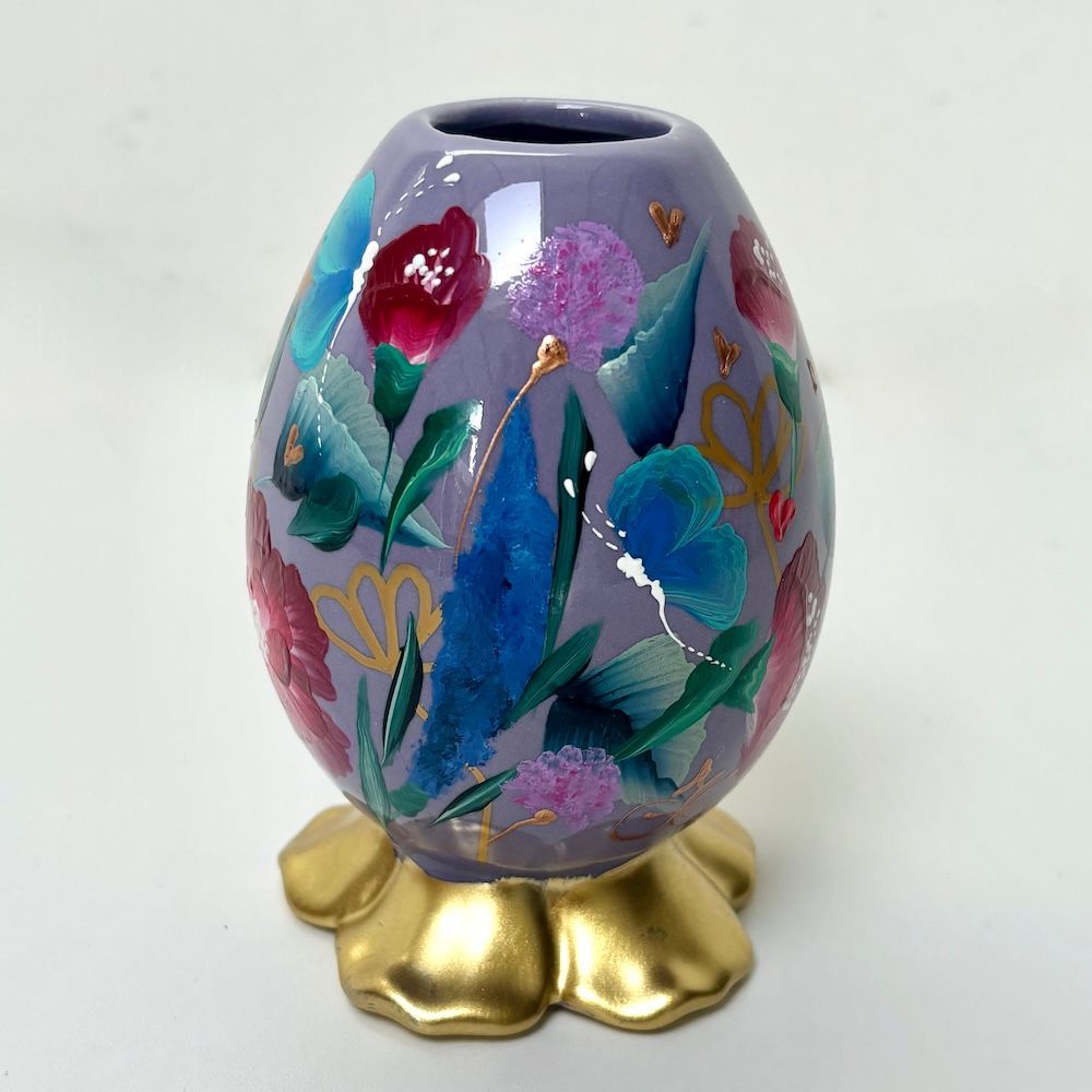 Een lavendelkleurige, eivormige vaas met handgeschilderde, kleurrijke bloemen op een gouden, vijfbladige bloemvoet.