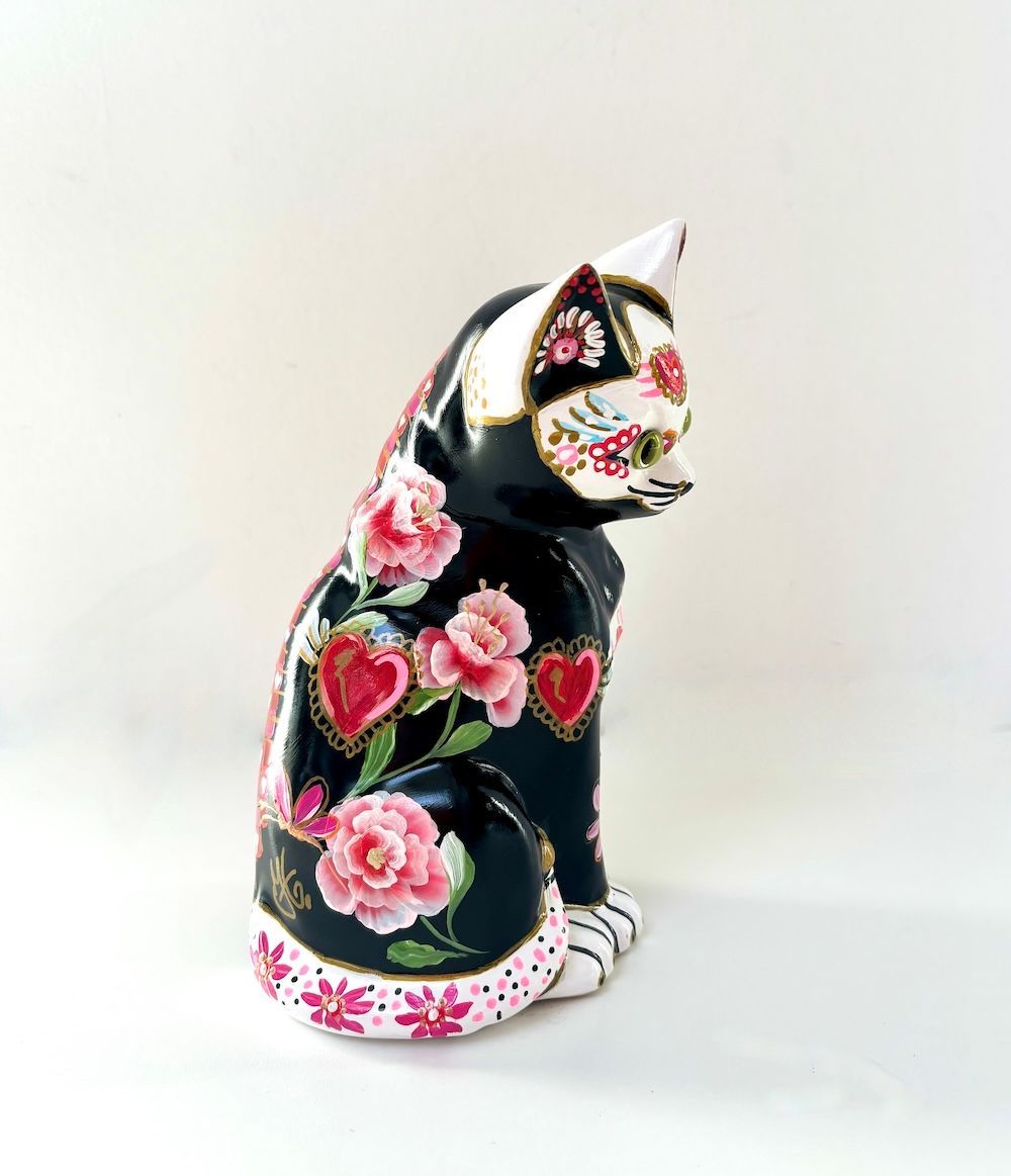 Een beeldje van een zwarte kat, versierd in de stijl van Día de Muertos ,waaronder roze hartjes, bloemen en witte details.