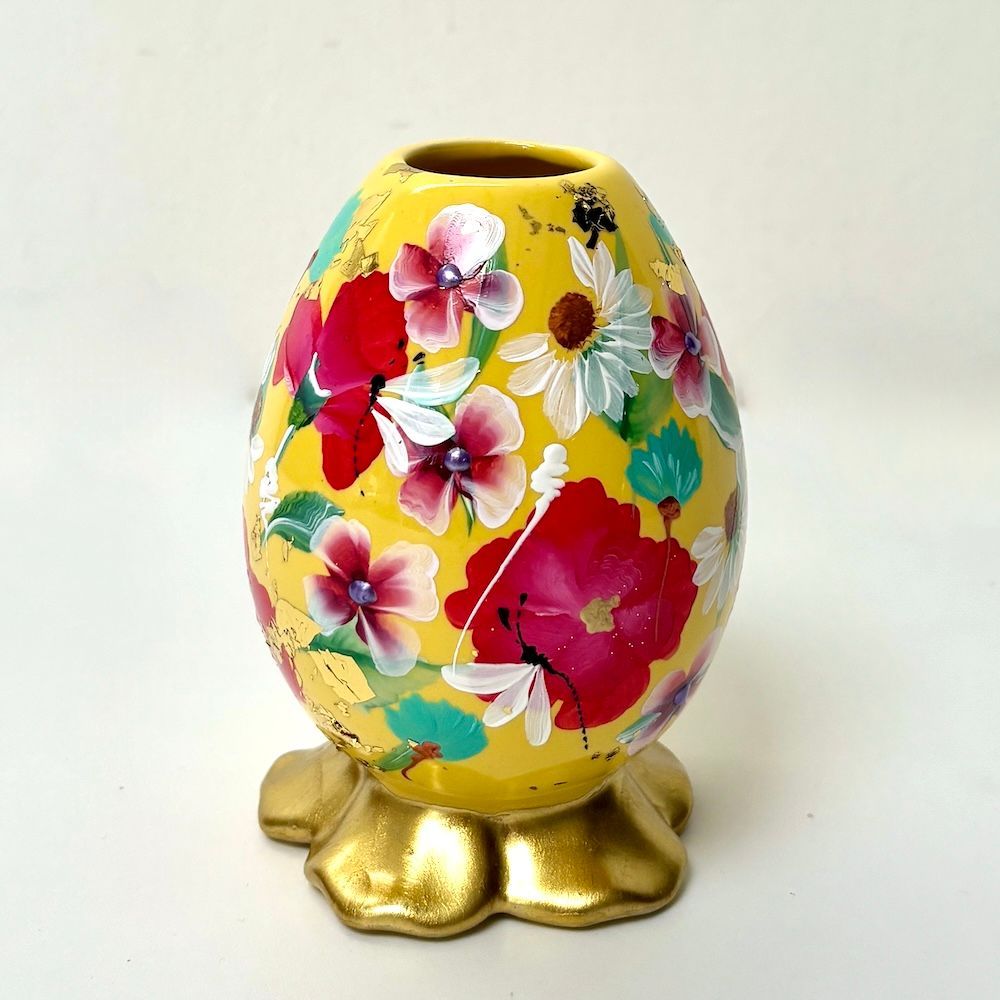 Een geel, eivormig vaasje, beschilderd met rode, witte en roze bloemen, staat op een gegolfde, metallic gouden voet.