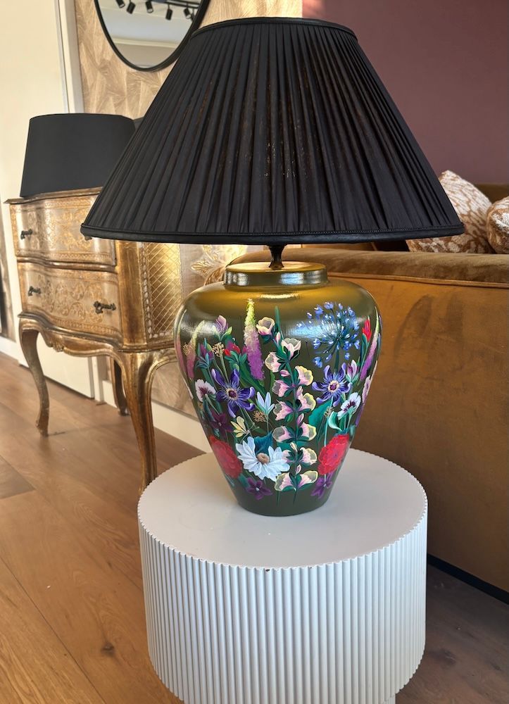 Decoratieve tafellamp met bloemmotief en zwarte geplooide kap op een donkere tafel tegen de muur.