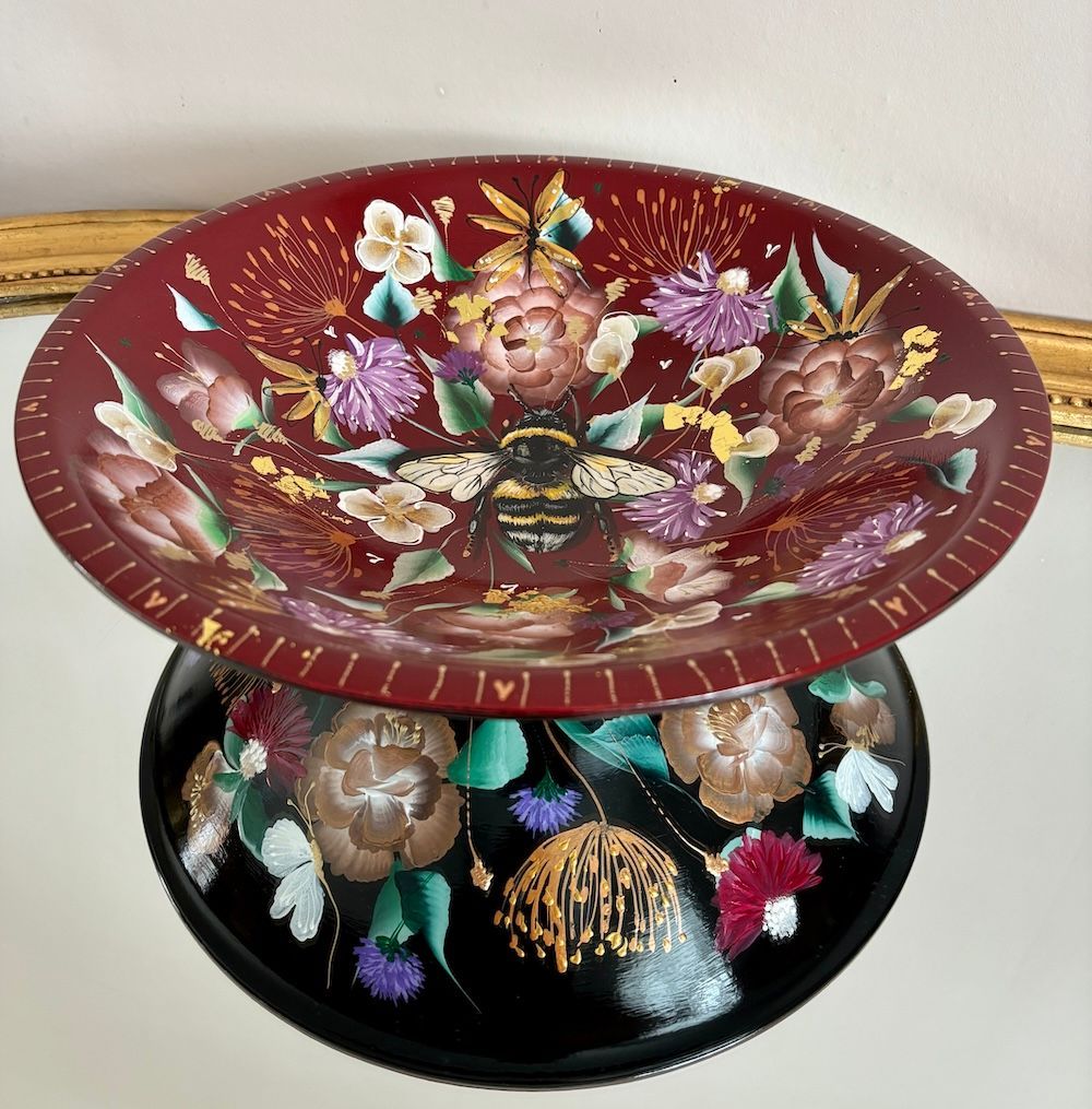 Een decoratieve schaal  met een dieprode binnenkant , beide handbeschilderd met kleurrijke bloemmotieven en een bij.