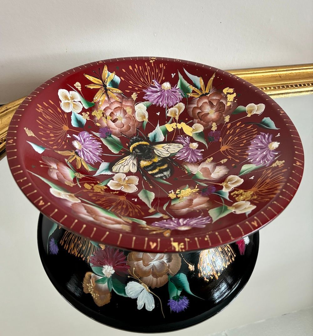 Een decoratieve schaal  met een dieprode binnenkant , beide handbeschilderd met kleurrijke bloemmotieven en een bij.