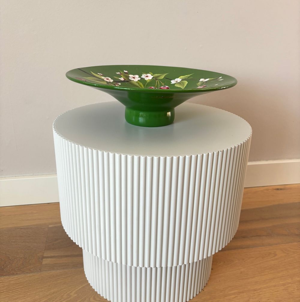 Een ronde, groene, handbeschilderde schaal met kersenbloesems en rode kersen staat op een witte, geplooide ronde tafel.