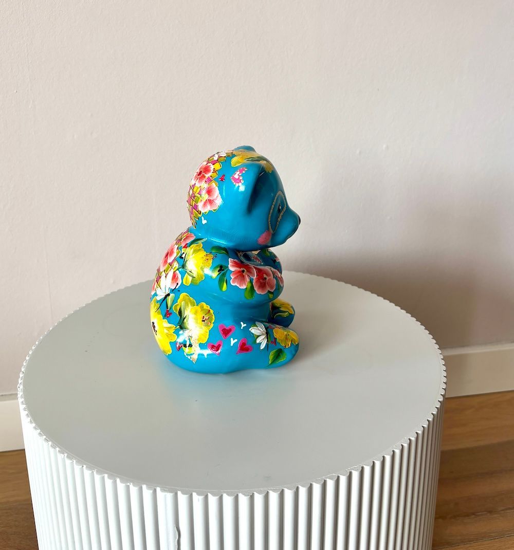 Een blauwe keramische beren beeld, beschilderd met bloemmotieven en hartjes, staat op een witte, ronde tafel met ribbels.
