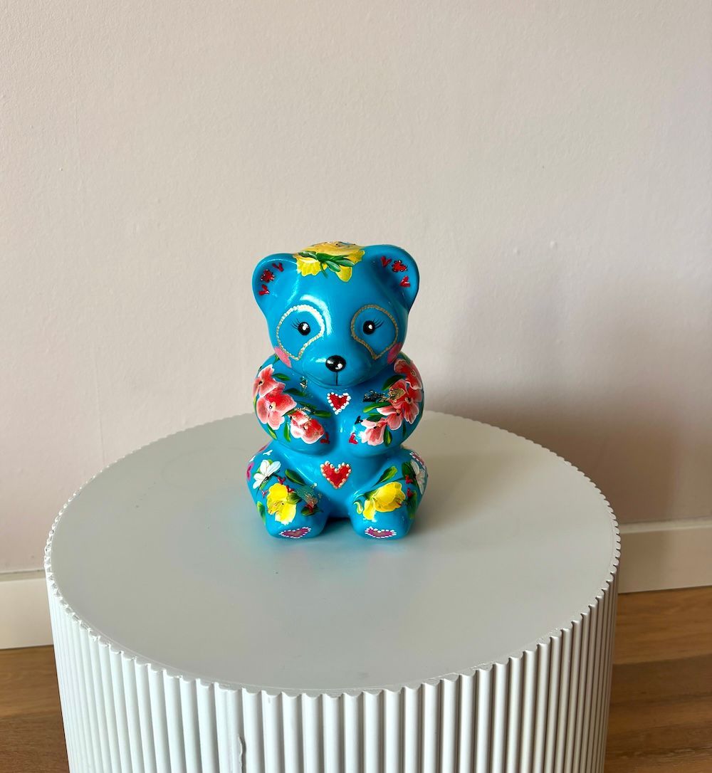 Een blauwe keramische beren beeld, beschilderd met bloemmotieven en hartjes, staat op een witte, ronde tafel met ribbels.