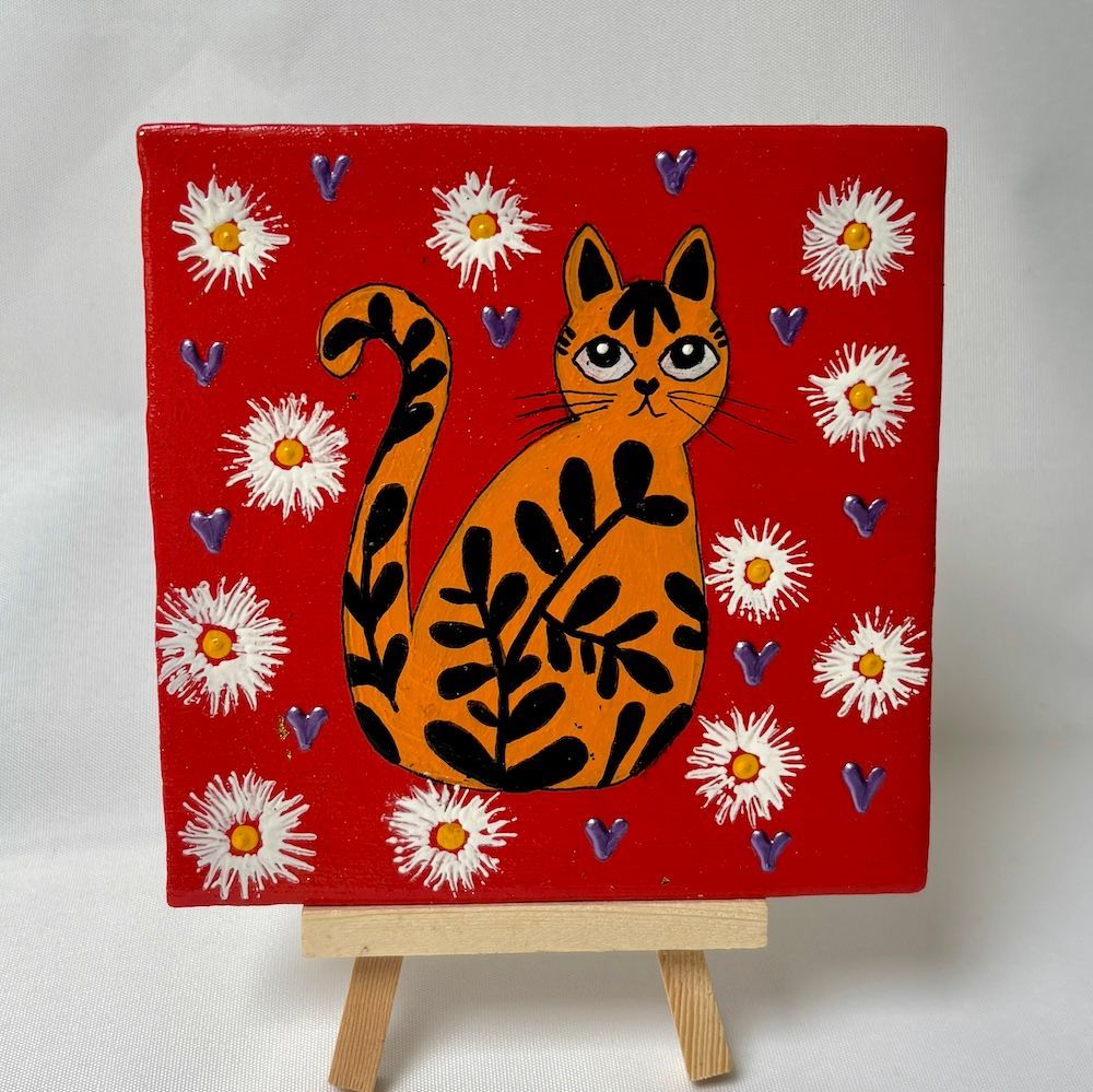 Een schilderij op rood canvas van een oranje kat met zwarte bladeren, omringd door witte bloemen, op een kleine schildersezel.