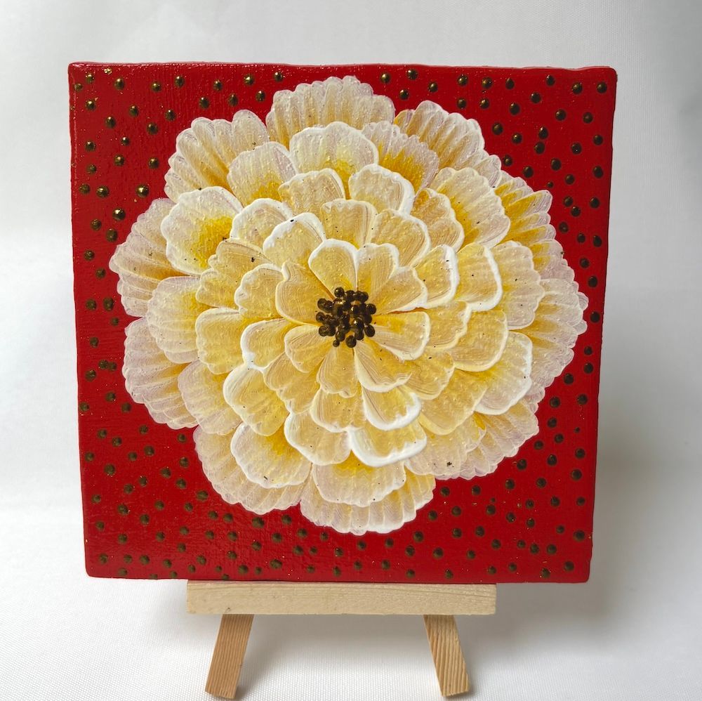 Een rood vierkant kunstwerk met een grote witte en gele bloem op een houten schildersezel.