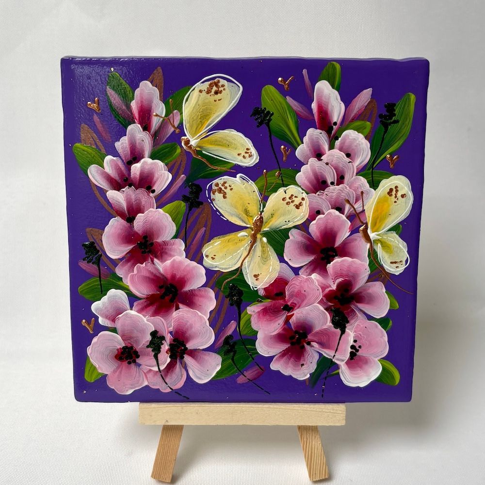 Op een klein, vierkant tegeltje zijn roze bloemen en lichtgele vlinders afgebeeld tegen een levendige paarse achtergrond.