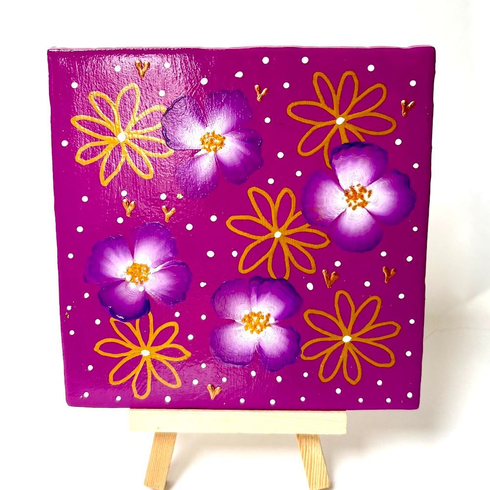 Paars bloemenschilderij op een kleine ezel met gouden en witte bloemen en verspreide glitters.