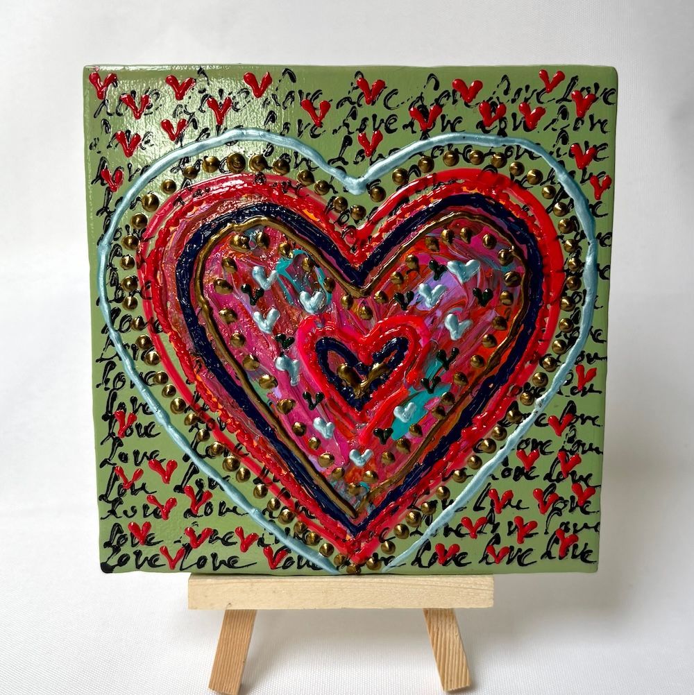Abstracte hartenschilderij op een kleine ezel, met gelaagde rode, roze en zwarte harten op een groene, gedessineerde achtergrond.
