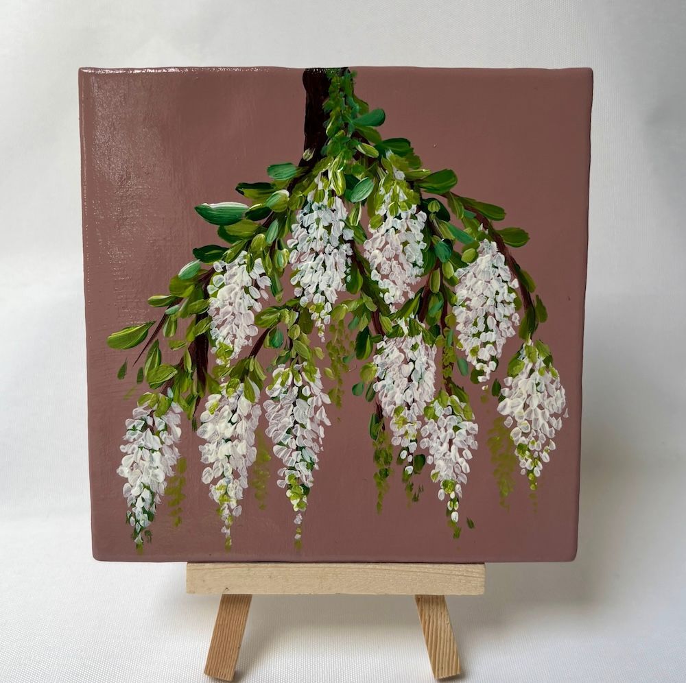 Witte blauweregenbloemen met groene bladeren geschilderd op een mauvekleurig vierkant doek op een kleine schildersezel.