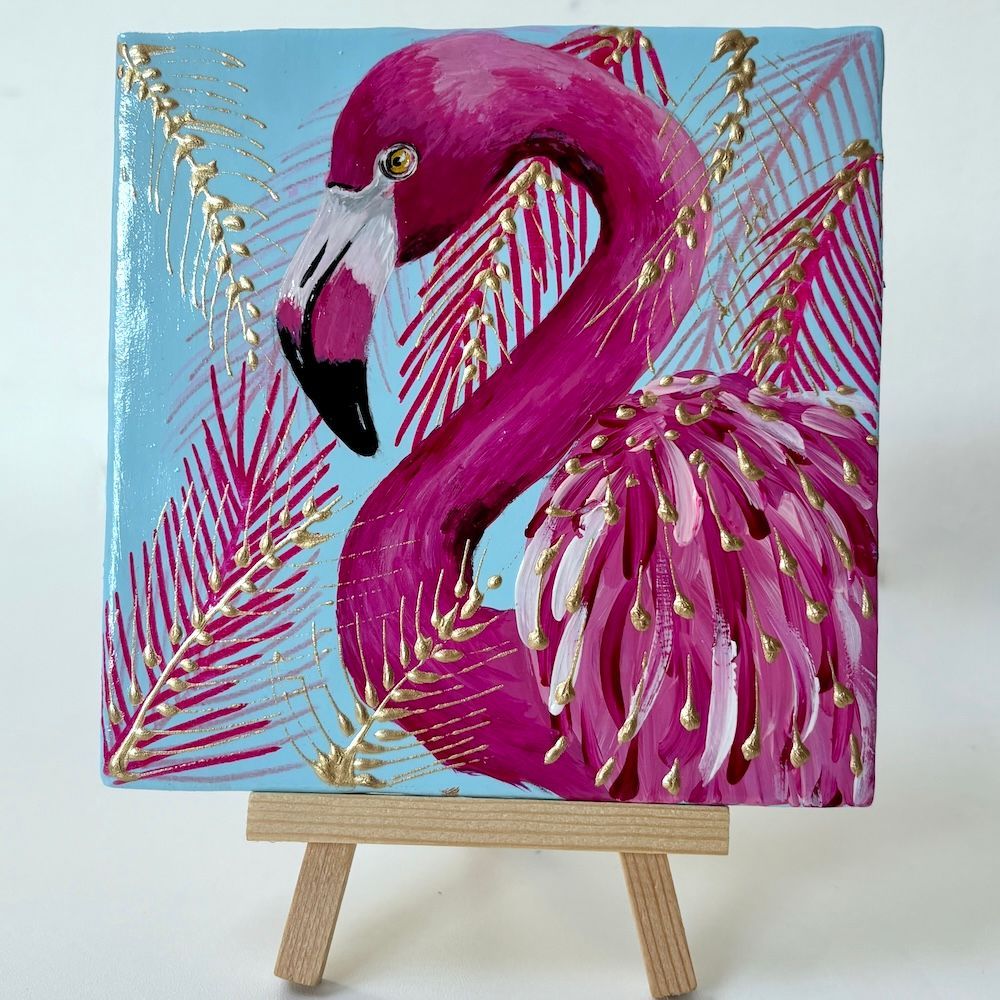 Schilderij van een roze flamingo op een kleine schildersezel, met gouden en roze palmbladeren op een blauwe achtergrond.