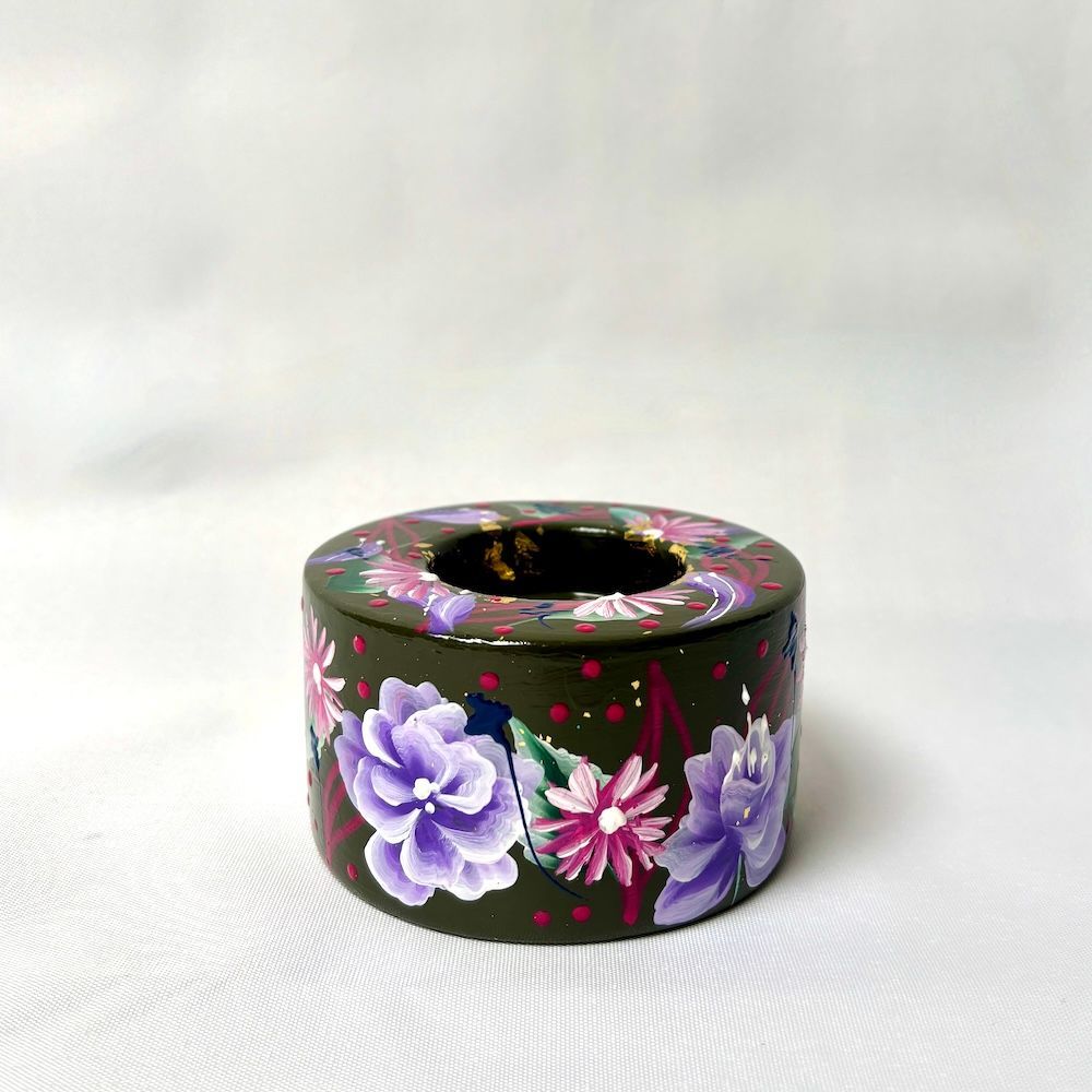 Een ronde, donkergroene houten kaarsenhouder, handbeschilderd met paarse en roze bloemenpatronen op een witte achtergrond.
