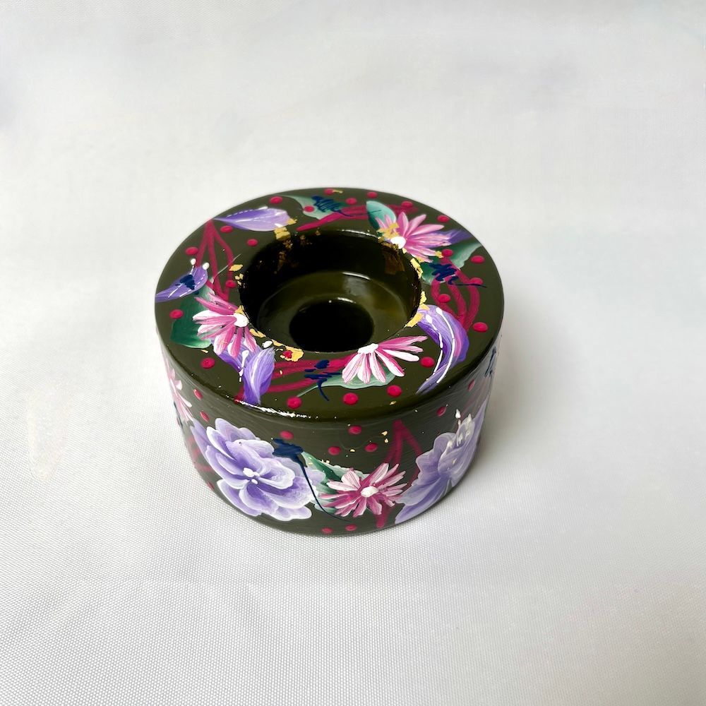 Een ronde, donkergroene houten kaarsenhouder, handbeschilderd met paarse en roze bloemenpatronen op een witte achtergrond.