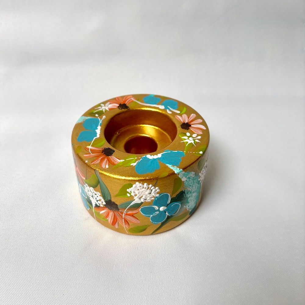 Een goudkleurige kaarsenhouder, versierd met handgeschilderde blauwe, witte en oranje bloemen op een witte achtergrond.