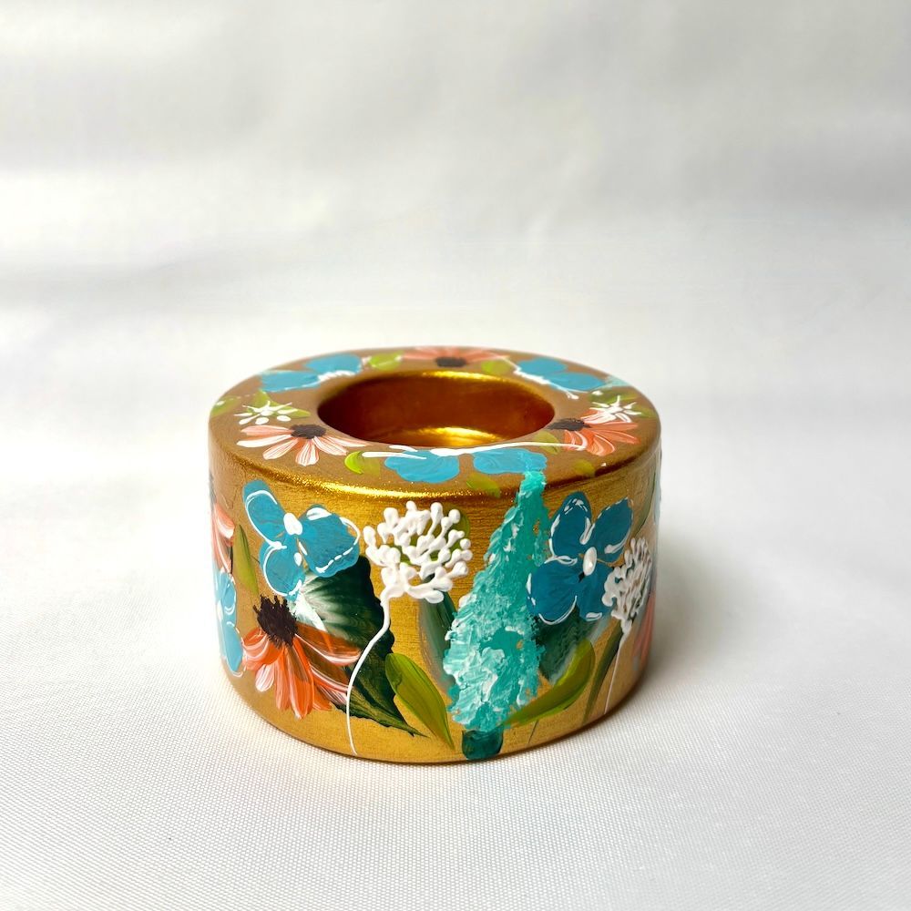 Een goudkleurige kaarsenhouder, versierd met handgeschilderde blauwe, witte en oranje bloemen op een witte achtergrond.