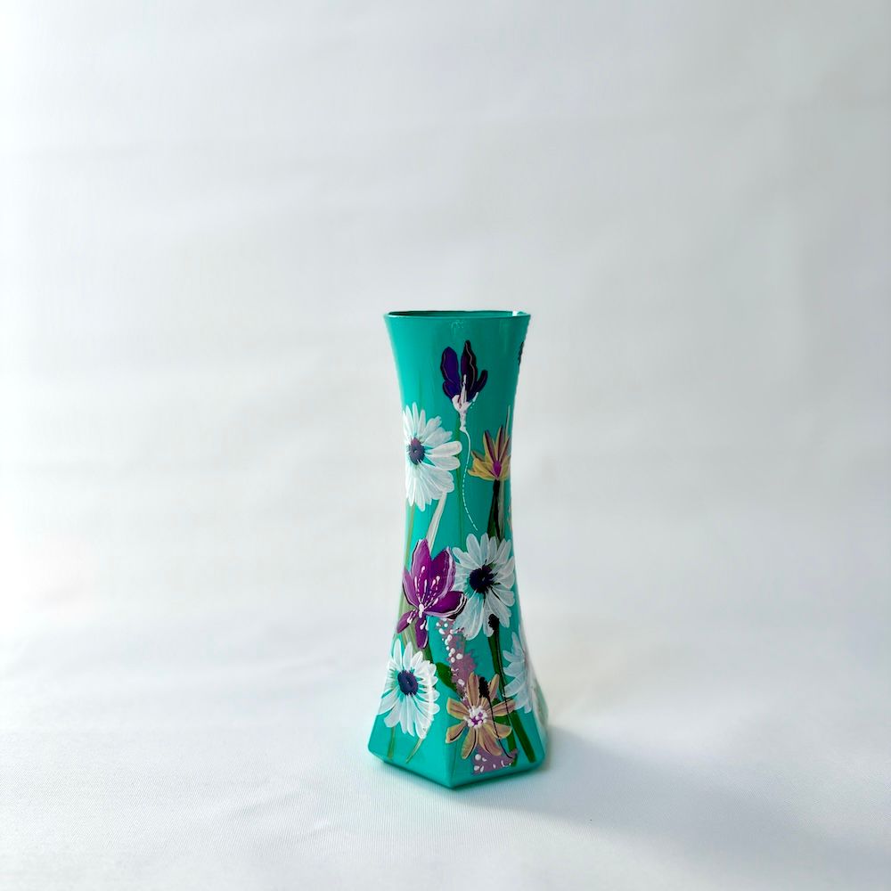 Een turquoise, zeshoekige vaas, handbeschilderd met witte en paarse bloemen, geplaatst tegen een effen witte achtergrond.