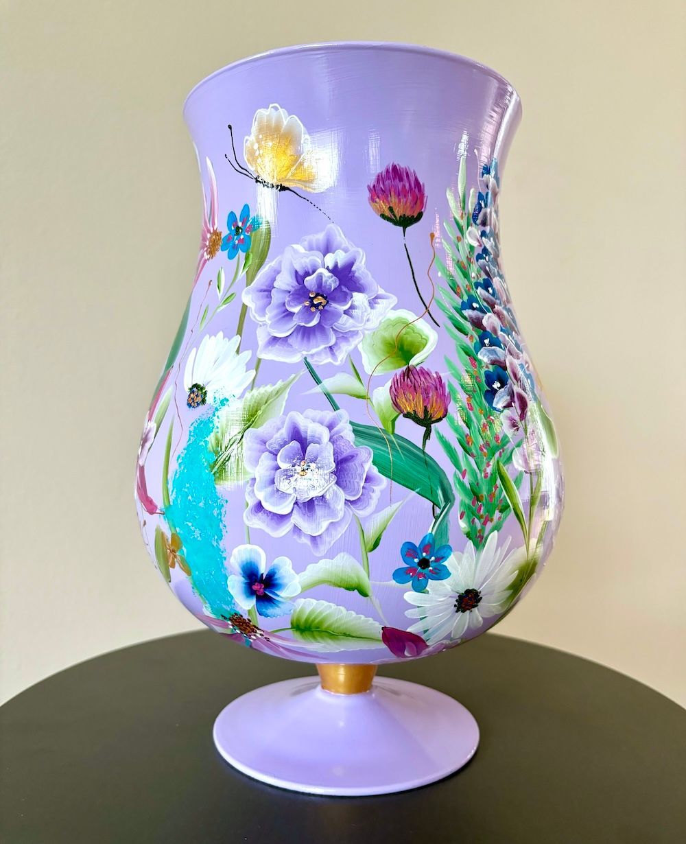 Een handgeschilderde vaas op een voetstuk, lila beschilderd met kleurrijke, gedetailleerde bloemen en een kleine vlinder.