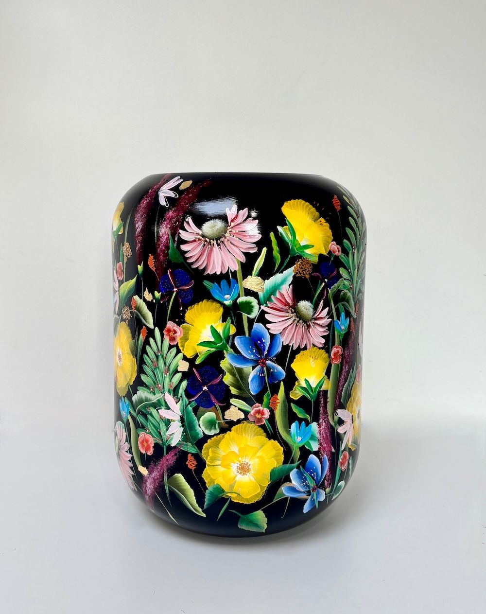 Een cilindrische zwarte vaas versierd met een levendig, kleurrijk schilderij van diverse tuinbloemen en groen gebladerte.