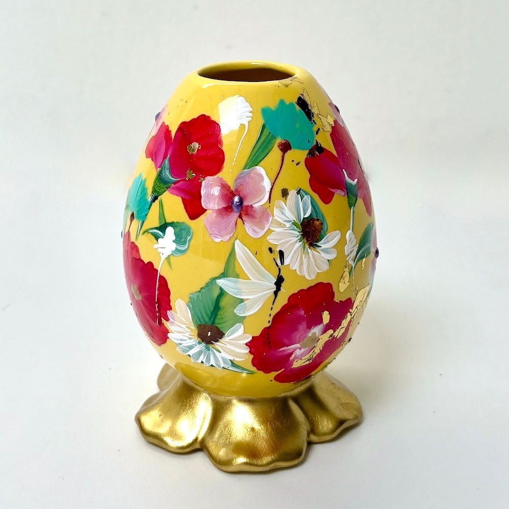 Een geel, eivormig vaasje, beschilderd met rode, witte en roze bloemen, staat op een gegolfde, metallic gouden voet.