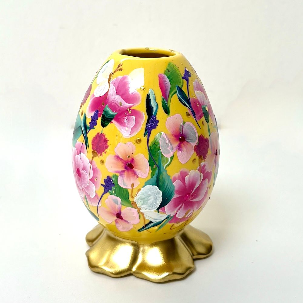 Een gele, eivormige vaas versierd met geschilderde roze en witte bloemen rustend op een gouden, geschulpte voet.