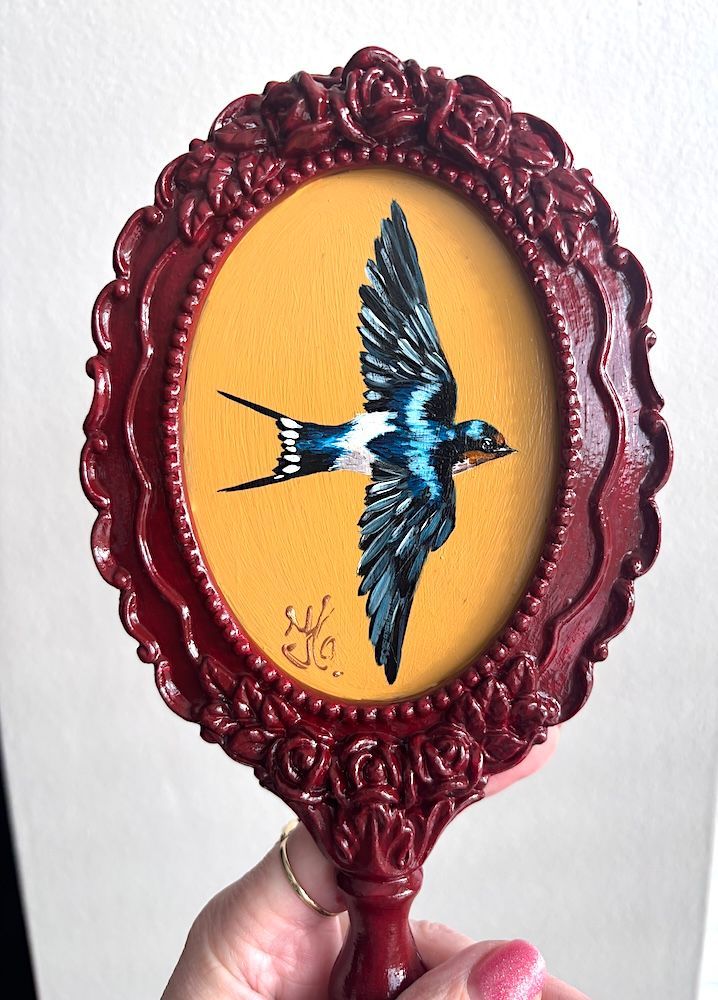 Sierlijke rode handspiegel met een ovaal gouden middenstuk waarop een blauw vogelsilhouet is afgebeeld.